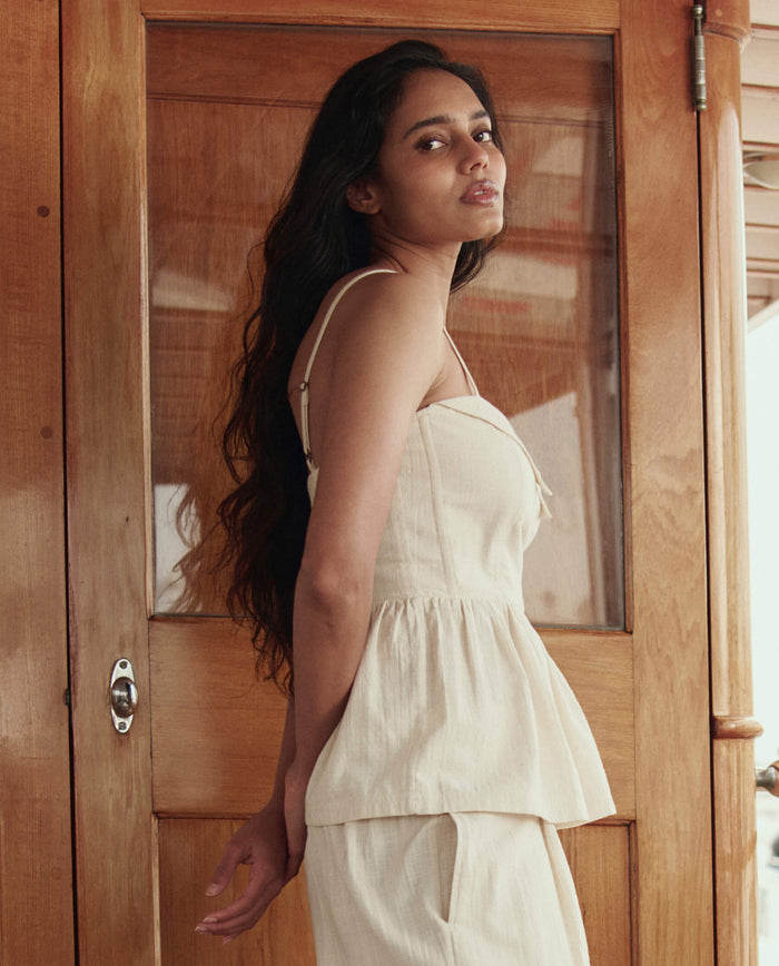 A woman with long dark hair wears THE GREAT. SU26's The Verse Top in Natural, paired with light pants, as she stands before a wooden door and looks over her shoulder.