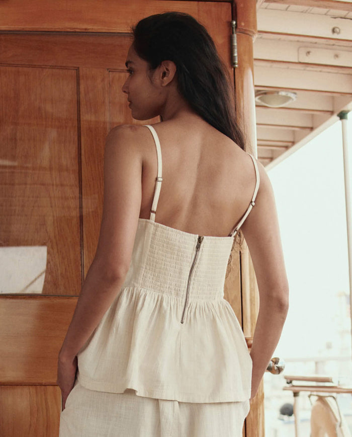 A person with long hair stands indoors, facing away, wearing THE GREAT. SU26’s The Verse Top in Natural—a cream-colored, sleeveless top with thin straps and a back zipper.