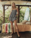 A woman wears THE GREAT. SP26 SWIM's The Tide Smocked Top in Black & Cream Check with cowboy boots, standing indoors by a bed with a striped blanket; a surfboard is visible in the background.