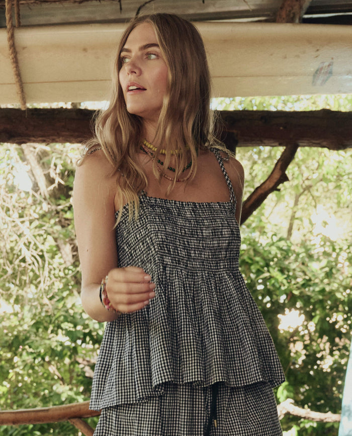 A woman with long, wavy hair wears THE GREAT. SP26 SWIM's The Tide Smocked Top in Black & Cream Check, paired with beaded necklaces, standing outdoors near wooden beams and greenery.