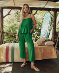 A woman stands indoors by a bed, wearing THE GREAT. SP26 SWIM's The Tide Smocked Top in Bright Sprig with matching pants and accessories. Surfboards and trees can be seen in the background.