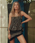 A woman with long hair poses by surfboards, wearing THE GREAT. SP26 SWIM’s The Tide Smocked Top in Leopard—a stylish snapshot of women’s fashion.