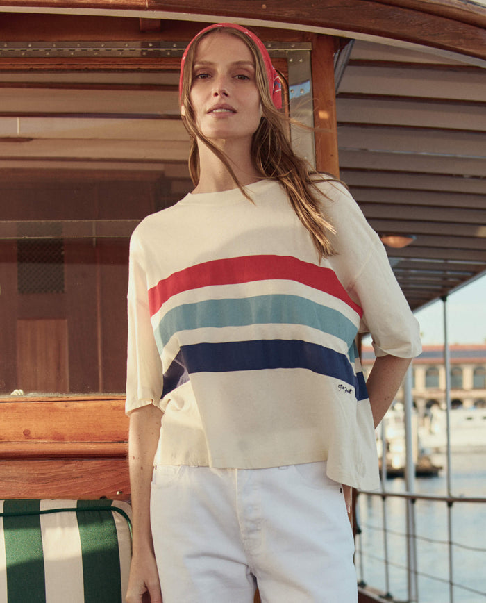 A woman stands on a boat with wooden details and a marina behind her, wearing THE GREAT. SU26 The Track Crew W/ Saltwater Stripe in Washed White and white pants.