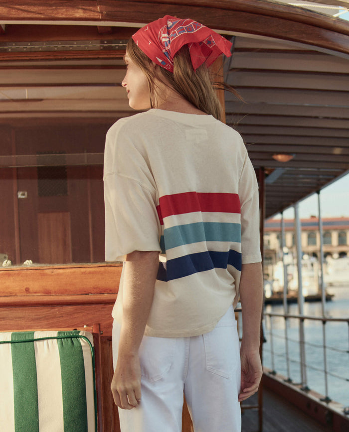 A woman wearing THE GREAT. SU26 The Track Crew W/ Saltwater Stripe in Washed White and white pants stands on a boat, facing away, with a red scarf tied in her hair.