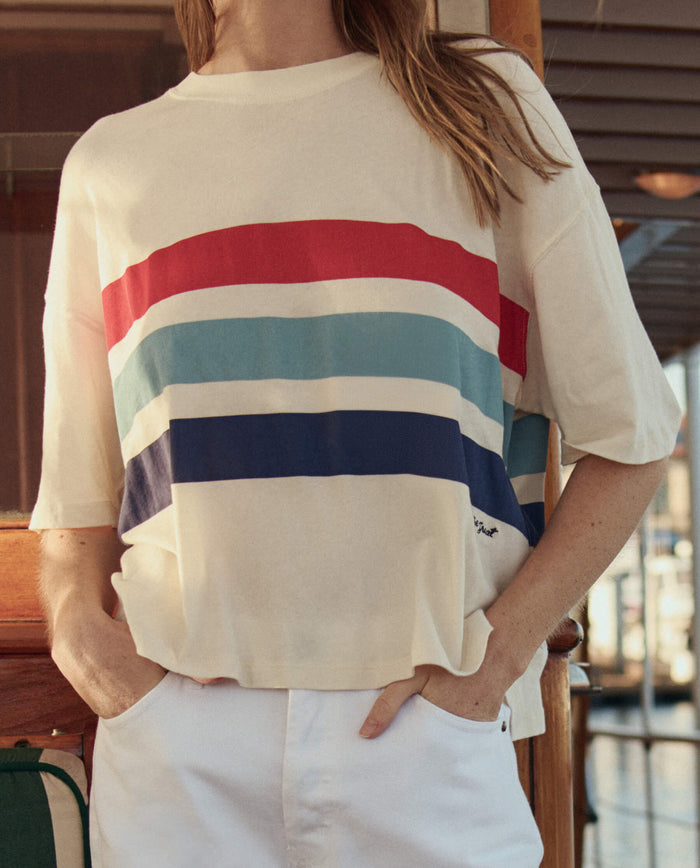 Someone wearing The Track Crew W/ Saltwater Stripe in Washed White by THE GREAT. SU26—a cream-colored top with red, light blue, teal, and navy stripes—paired with white pants.