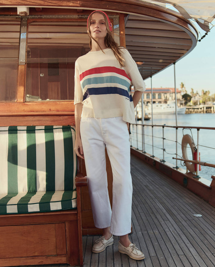 A woman stands on a wooden boat deck wearing THE GREAT. SU26 The Track Crew W/ Saltwater Stripe in Washed White, white pants, tan shoes, and a red headband. A striped bench and marina form the backdrop.
