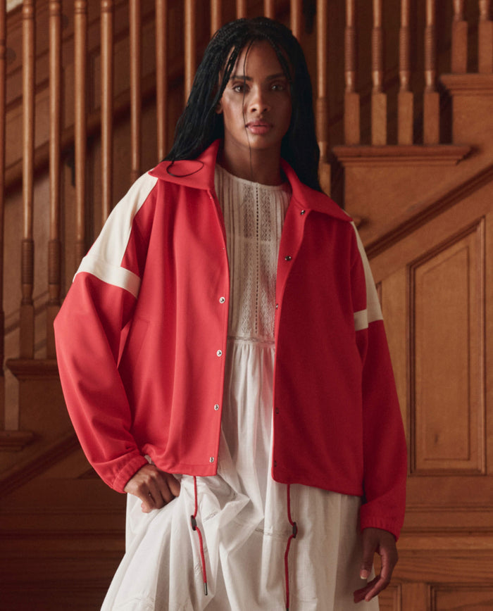 A person stands indoors before wooden stairs, wearing THE GREAT. SP26 D1 The Trainer Jacket in Poppy Red over a white dress.
