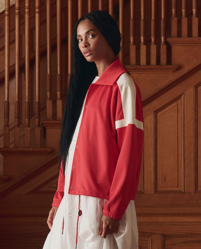 A woman with long black hair wears THE GREAT. SP26 D1 The Trainer Jacket in Poppy Red over a white dress, standing before a wooden staircase.