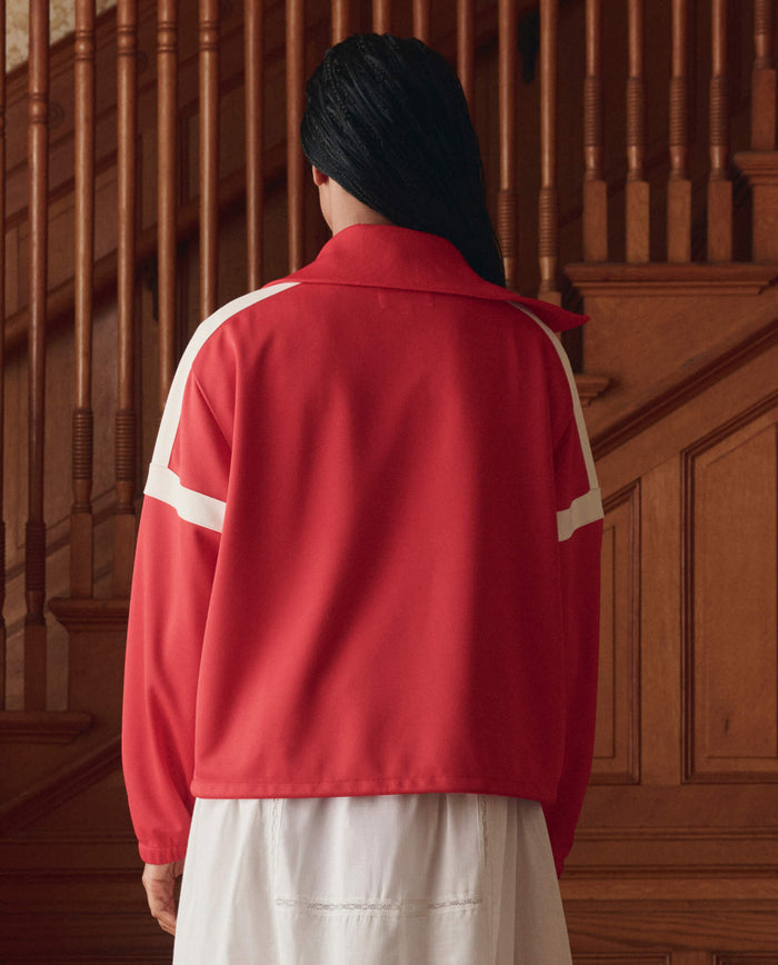 A person with long dark hair wears THE GREAT. SP26 D1 The Trainer Jacket in Poppy Red over a white dress, standing indoors facing away from the camera in front of a wooden staircase.