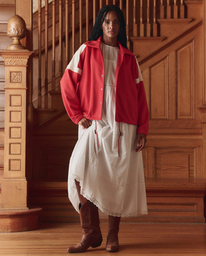 A person stands indoors on wooden flooring, wearing a white dress, brown cowboy boots, and THE GREAT. SP26 D1 The Trainer Jacket in Poppy Red, with a wooden staircase in the background.
