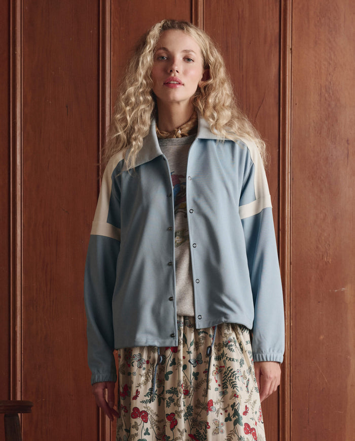 A woman with long curly hair stands before a wooden paneled wall, wearing THE GREAT. SP26 D1's The Trainer Jacket in Robin Blue over a gray shirt and a floral skirt.