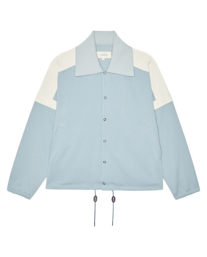 The Trainer Jacket by THE GREAT. SP26 D1 in Robin Blue features snap buttons, side pockets, drawstring hem, and elastic cuffs, shown on a plain white background.