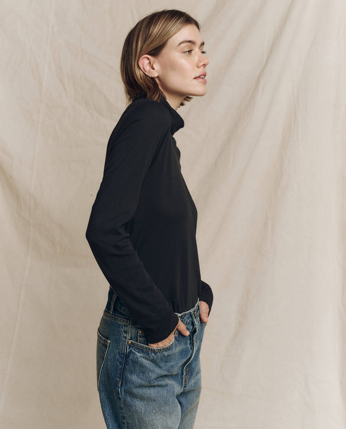 A model wearing The Turtleneck in Almost Black from THE GREAT. HOL 24 TURTLENECKS pairs it with jeans and stands sideways against a beige fabric backdrop, embodying timeless fashion with minimalist elegance.