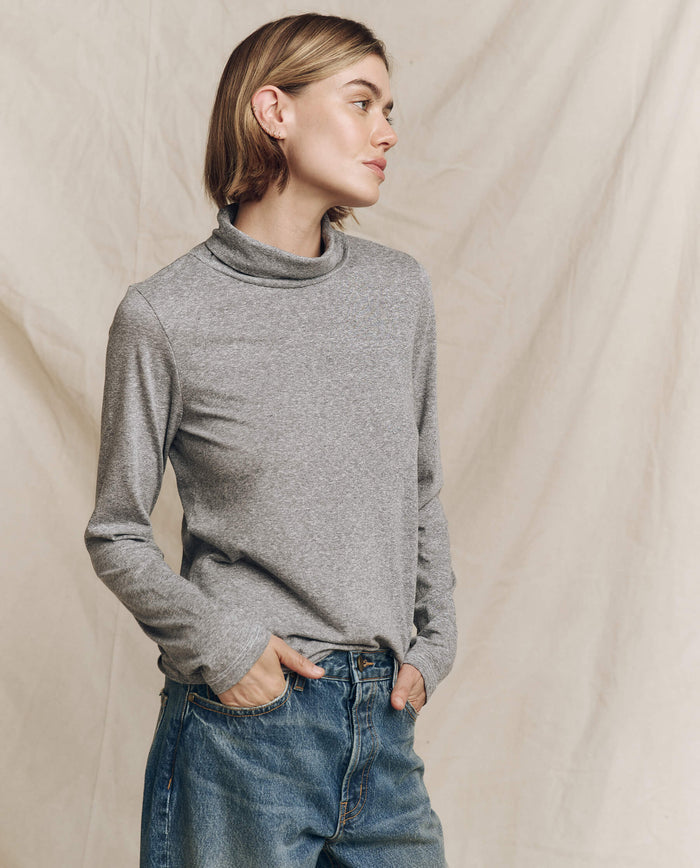 A person wearing THE GREAT. HOL 24 TURTLENECKS' "The Turtleneck" in Heather Grey and blue jeans stands against a beige backdrop, gazing to the side.