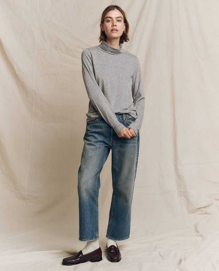 A person wearing The Turtleneck in Heather Grey from THE GREAT. HOL 24 TURTLENECKS and blue jeans stands against a beige backdrop.