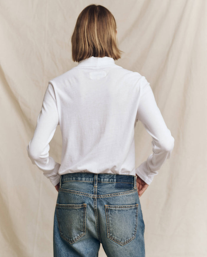 A person with shoulder-length hair is wearing "The Turtleneck" in True White by THE GREAT. HOL 24 TURTLENECKS, along with blue jeans, standing with their back to the camera in front of a beige backdrop.
