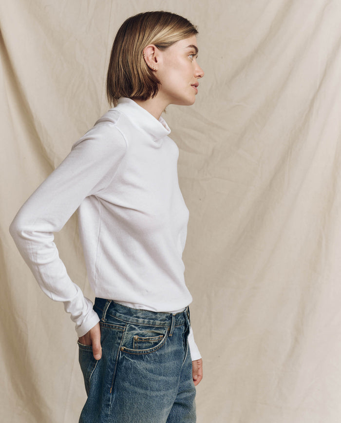 A figure in "The Turtleneck" by THE GREAT. HOL 24 TURTLENECKS, in True White, and jeans stands poised in profile against a soft beige fabric backdrop, effortlessly blending minimalist style with timeless elegance.