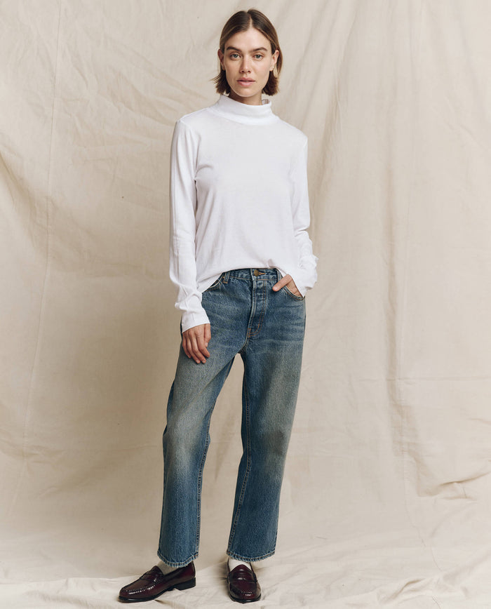 A person wearing "The Turtleneck" in True White from THE GREAT. HOL 24 TURTLENECKS, along with blue jeans and dark loafers, stands against a beige backdrop.