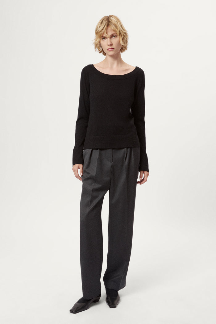Person wearing a black long-sleeve top and dark gray pleated pants on a white background