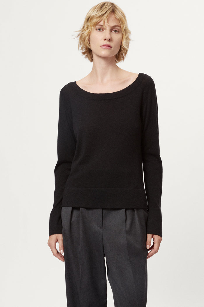 Person wearing a black long-sleeve top and dark gray pleated pants on a white background