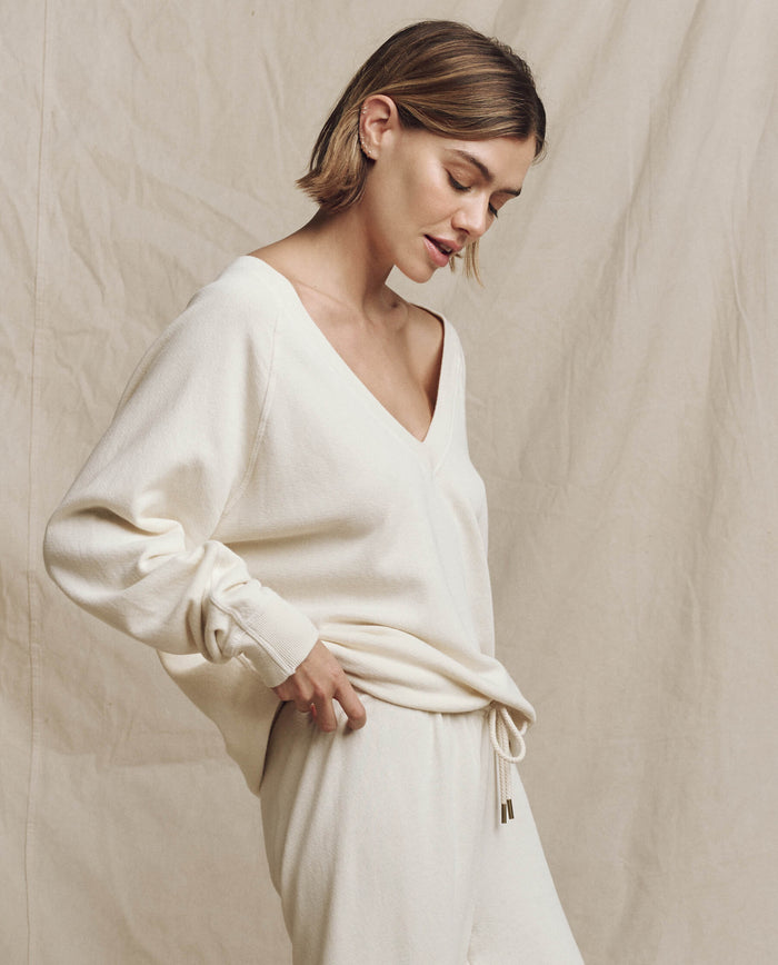 A woman with short hair wearing The V-Neck Sweatshirt in washed white and matching pants from THE GREAT. FALL 24 KNITS stands against a beige background, looking down with her hand on her hip.