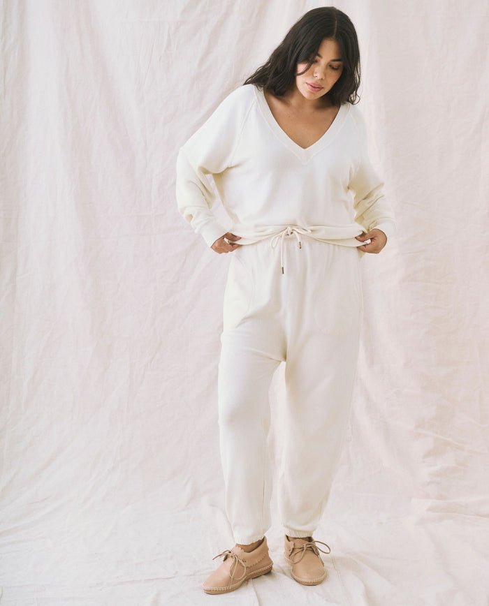 A woman stands against a light-colored backdrop, wearing The V-Neck Sweatshirt in Washed White by THE GREAT. FALL 24 KNITS and matching pants with a drawstring at the waist. She is looking down, with her hands adjusting the drawstring.