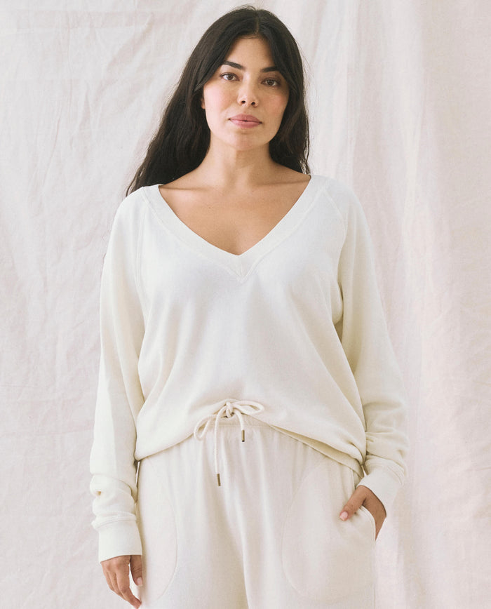 A person with long dark hair wears "The V-Neck Sweatshirt" in Washed White by THE GREAT. FALL 24 KNITS, along with matching pants, standing against a light-colored, textured background.