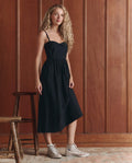 A woman with long curly hair wears THE GREAT. SP26 D1 The Verse Dress in black and white sneakers, standing indoors by wood-paneled walls near a wooden chair.