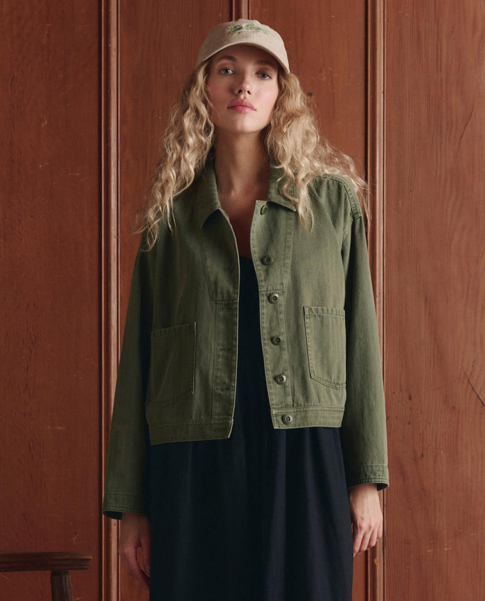 A woman with long, wavy blonde hair wears a beige baseball cap, an olive green jacket, and THE GREAT. SP26 D1 The Verse Dress in Black while standing in front of a wooden wall.