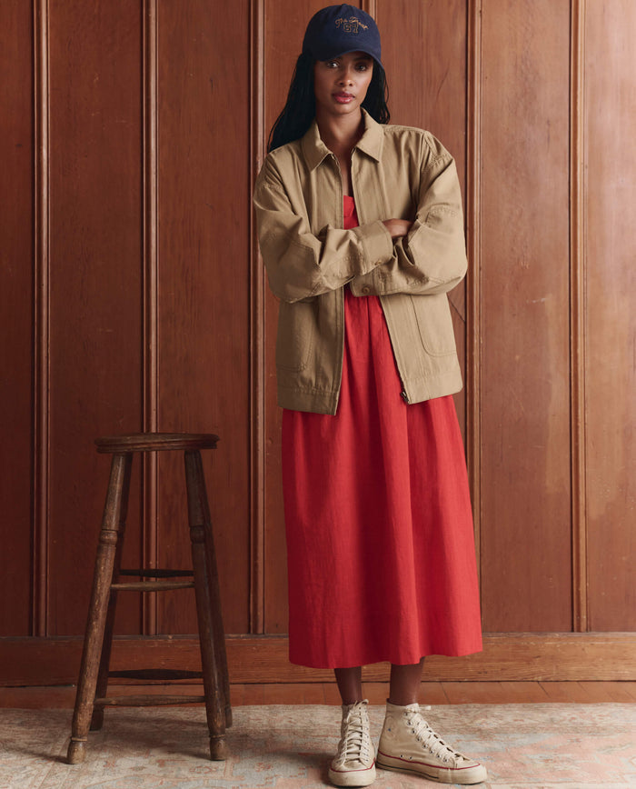 A woman stands indoors next to a wooden stool, wearing THE GREAT. SP26 D1 The Verse Dress in Hot Red with a sweetheart neckline, layered under a tan jacket, paired with white high-top sneakers and a navy cap.