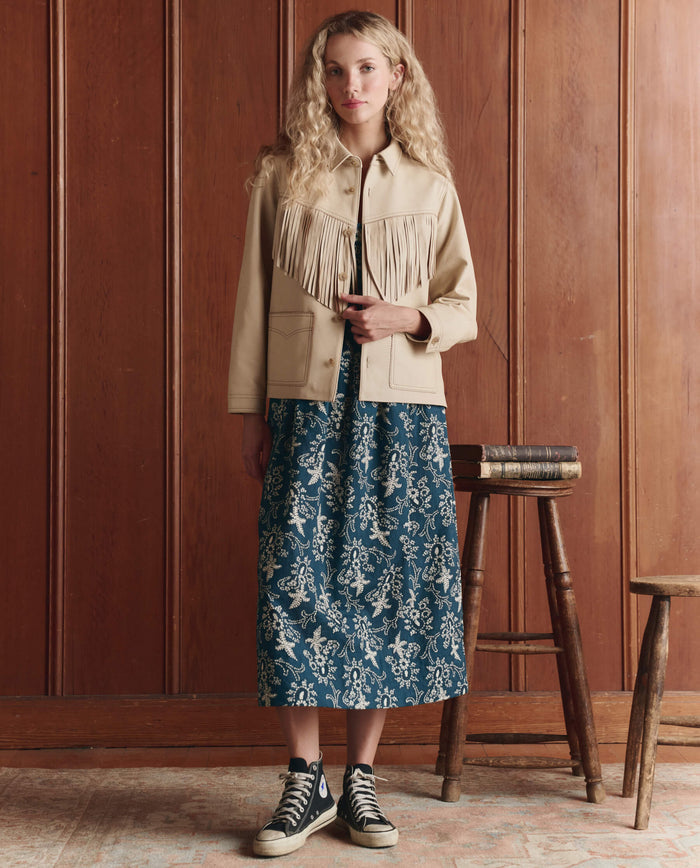 A woman stands indoors against wooden paneling, wearing THE GREAT. SP26 D1 The Leather Fringe Jacket in Toasted Almond, a blue patterned midi skirt, and black high-top sneakers. A stack of books sits on a small table beside her.