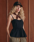 A woman with long, wavy blonde hair wears THE GREAT. SP26 D1 The Verse Top in black, striped pants, a brown neck scarf, and a black baseball cap while standing in front of a wooden paneled wall.