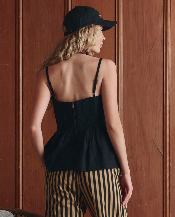 A person with long wavy hair, wearing THE GREAT. SP26 D1 The Verse Top in Black, a black cap, and black-and-yellow striped pants, stands with their back to the camera in front of a wooden wall.