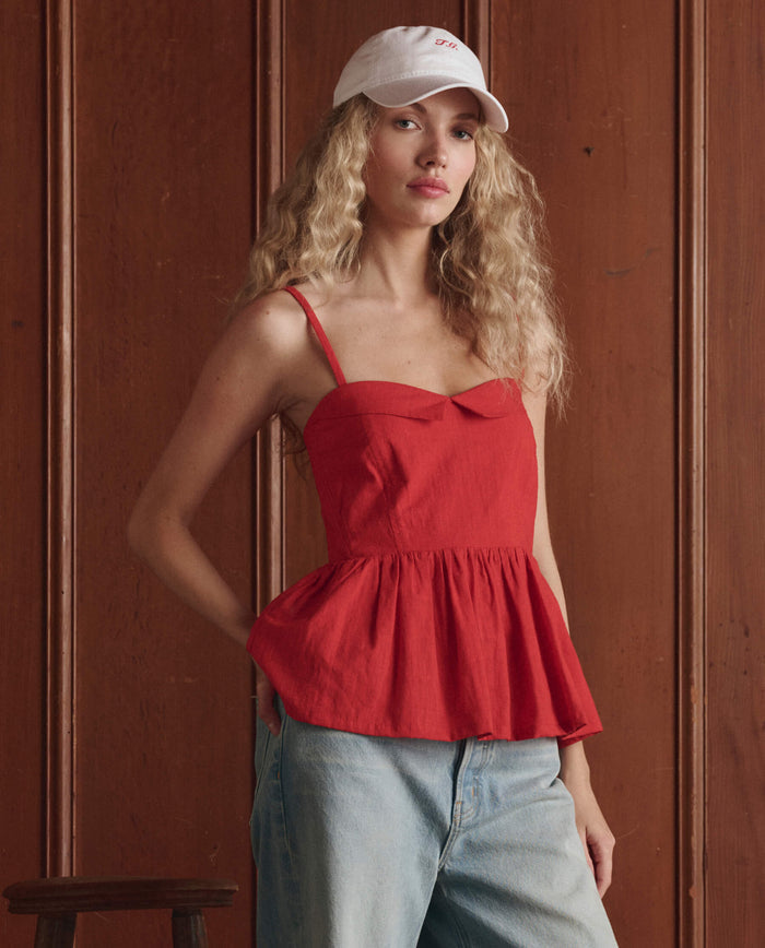 A woman stands before a wooden wall, wearing THE GREAT. SP26 D1’s The Verse Top in Hot Red—a sleeveless peplum women's blouse—paired with a white baseball cap and light blue jeans.