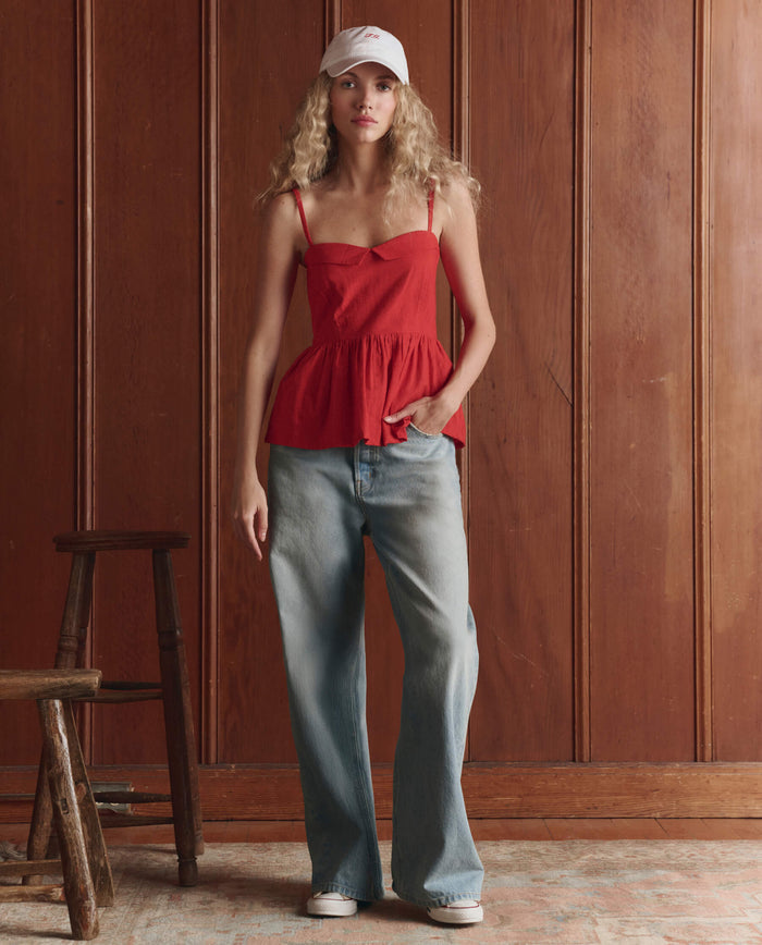 A woman in a white cap, loose light blue jeans, and THE GREAT. SP26 D1's The Verse Top -- Hot Red stands indoors by a wooden paneled wall.