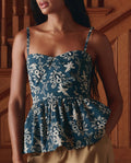 A woman stands indoors by a wooden staircase, wearing THE GREAT.'s The Verse Top in Indigo Bandana—featuring a blue and white floral pattern, sleeveless peplum style, sweetheart neckline, and thin straps.