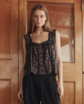 Indoors by wooden doors, a woman wears THE GREAT. SU26’s Victorian Pleat Tank in Black And Cream Token Floral with buttons, paired with loose black pants.