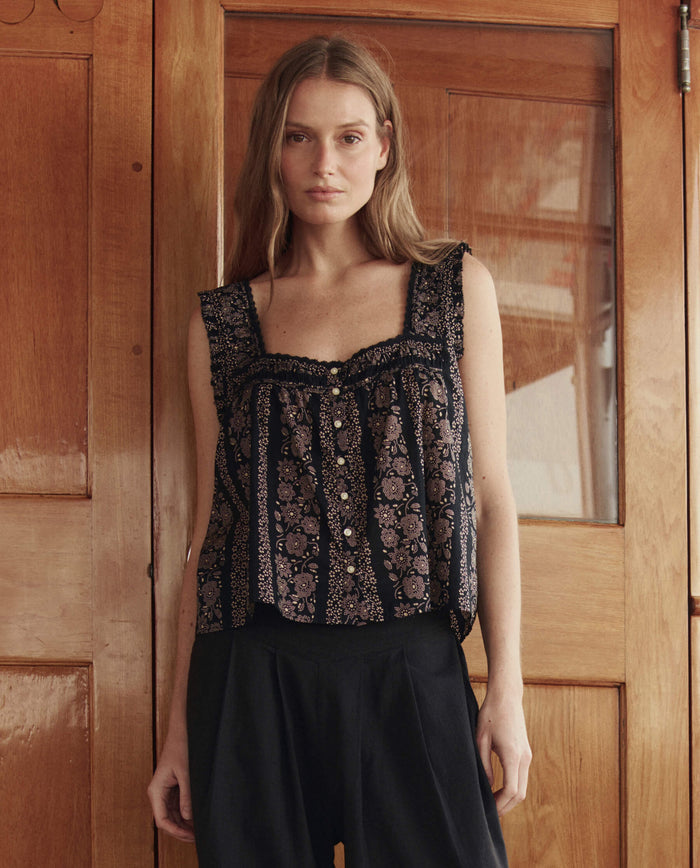 Indoors by wooden doors, a woman wears THE GREAT. SU26’s Victorian Pleat Tank in Black And Cream Token Floral with buttons, paired with loose black pants.