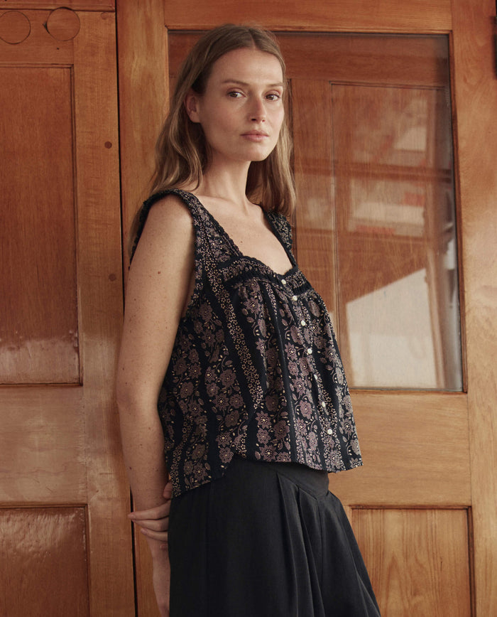 A woman with long hair stands indoors, turned slightly to the side, wearing THE GREAT. SU26 The Victorian Pleat Tank in Black and Cream Token Floral and dark pants, in front of wooden doors with glass panels.