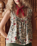 A woman with curly blonde hair wears THE GREAT. SP26 D1's The Victorian Pleat Tank in Floating Butterfly, paired with a red scarf, standing indoors beside carved wooden furniture.