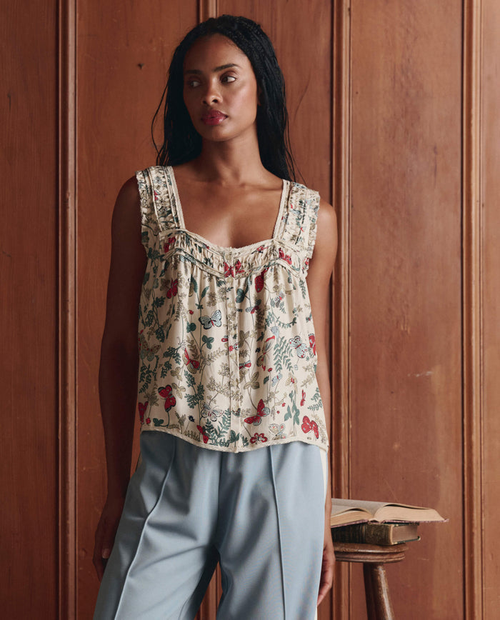 A woman wears THE GREAT. SP26 D1's The Victorian Pleat Tank -- Floating Butterfly with light blue pants, standing indoors by wooden paneling, an open book resting on a small stool nearby.