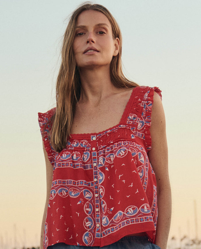 A woman with long hair wears THE GREAT. SU26's The Victorian Pleat Tank in Red Sailboat Bandana, featuring geometric and boat motifs, standing outdoors at sunset.