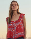 A woman with long hair wears THE GREAT. SU26's The Victorian Pleat Tank in Red Sailboat Bandana, featuring geometric and boat motifs, standing outdoors at sunset.