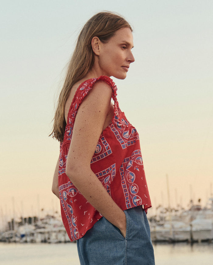 A woman wears THE GREAT.'s Victorian Pleat Tank and a Red Sailboat Bandana, standing outdoors in blue jeans with a marina and boats behind her at sunset.