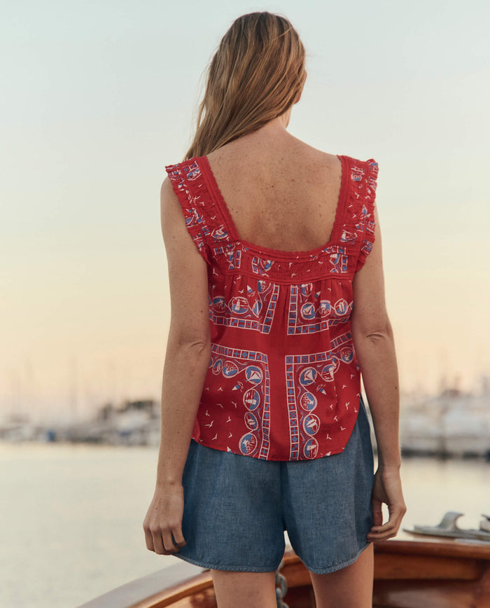 A woman with long hair stands on a boat, facing away, wearing THE GREAT. SU26 The Victorian Pleat Tank and the Red Sailboat Bandana, denim shorts, with a marina and boats in the background.