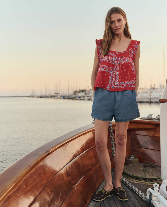 At sunset by the water, a woman stands on a wooden boat wearing THE GREAT. SU26 The Victorian Pleat Tank and a Red Sailboat Bandana. Denim shorts and boat shoes complete her look; sailboats and masts appear in the background.