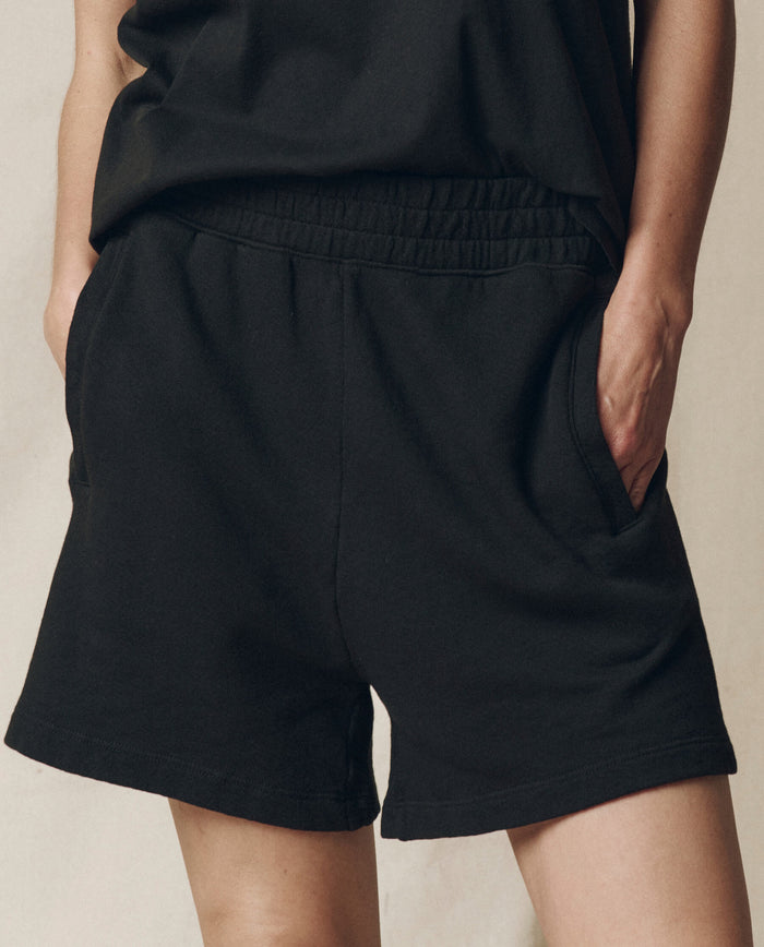 Model wears The Victory Short. Solid -- Almost Black by THE GREAT. SU25 CORE KNITS with a black top, standing hands-in-pockets against a neutral background and highlighting the shorts' comfortable, athletic fit.