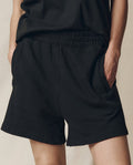 Model wears The Victory Short. Solid -- Almost Black by THE GREAT. SU25 CORE KNITS with a black top, standing hands-in-pockets against a neutral background and highlighting the shorts' comfortable, athletic fit.