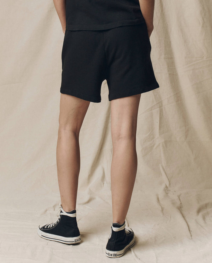 A person stands with their back to the camera wearing THE GREAT. SU25 CORE KNITS The Victory Short. Solid -- Almost Black, paired with a black shirt and high-top sneakers on a neutral fabric background, highlighting comfortable athletic wear.