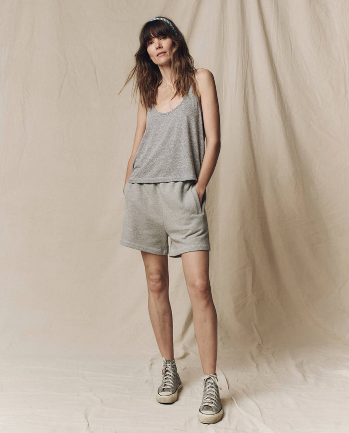 A woman stands with hands in pockets, wearing a gray tank top, THE GREAT. SU25 CORE KNITS The Victory Short. Solid -- Varsity Grey, and sneakers against a beige fabric backdrop.